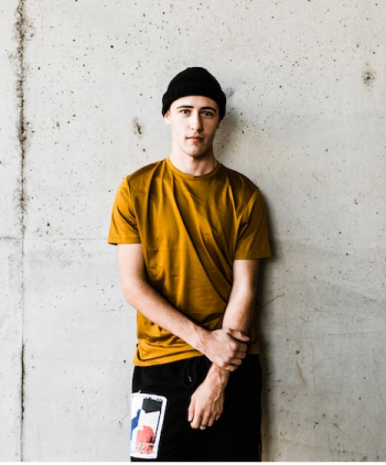 Modelo masculino vistiendo gorro negro, blusa marrón y pantalones jeans, con una pared gris de fondo