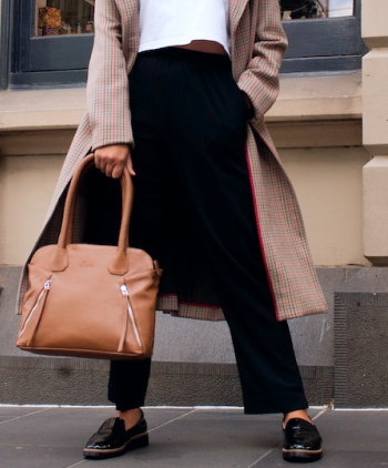 Cintura, piernas y pies de modelo femenina vistiendo abrigo de color predominante beige con detalles en tono rojo y oscuro, con pantalones y par de zapatos de color negro, sosteniendo una bolsa de color beige, con fondo de edificio