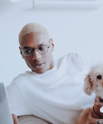 Modelo masculino con blusa blanca y un par de gafas de lentes transparentes redondeadas, utilizando un portátil, con un perro caniche blanco en su regazo, y en un fondo blanco