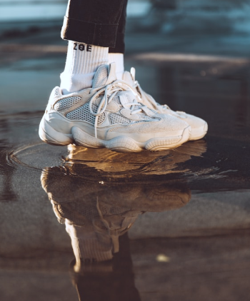 Enfoque en los pies de un modelo usando par de tenis y calcetines de color blanco, y pantalones de color negro, pisando en un charco de agua