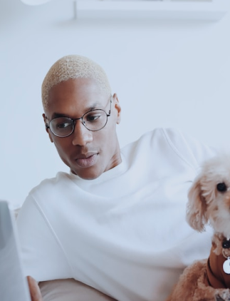 Modelo masculino con blusa blanca y un par de gafas de lentes transparentes redondeadas, utilizando un portátil, con un perro caniche blanco en su regazo, y en un fondo blanco