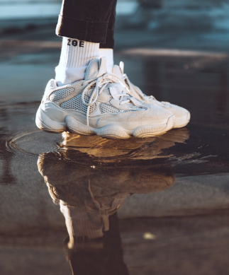 Enfoque en los pies de un modelo usando par de tenis y calcetines de color blanco, y pantalones de color negro, pisando en un charco de agua