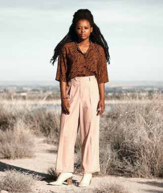 Modelo femenina vistiendo blusa marrón, pantalones beige, par de zapatos altos de color blanco, collar y pulsera, en un ambiente abierto con suelo de tierra y matorrales secos