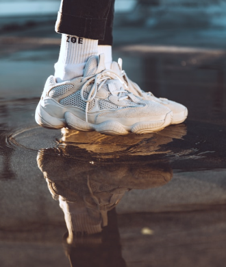 Enfoque en los pies de un modelo usando par de tenis y calcetines de color blanco, y pantalones de color negro, pisando en un charco de agua